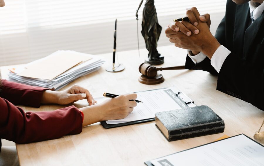 Two professionals at a desk reviewing and signing legal documents, with a gavel and a scale of justice nearby