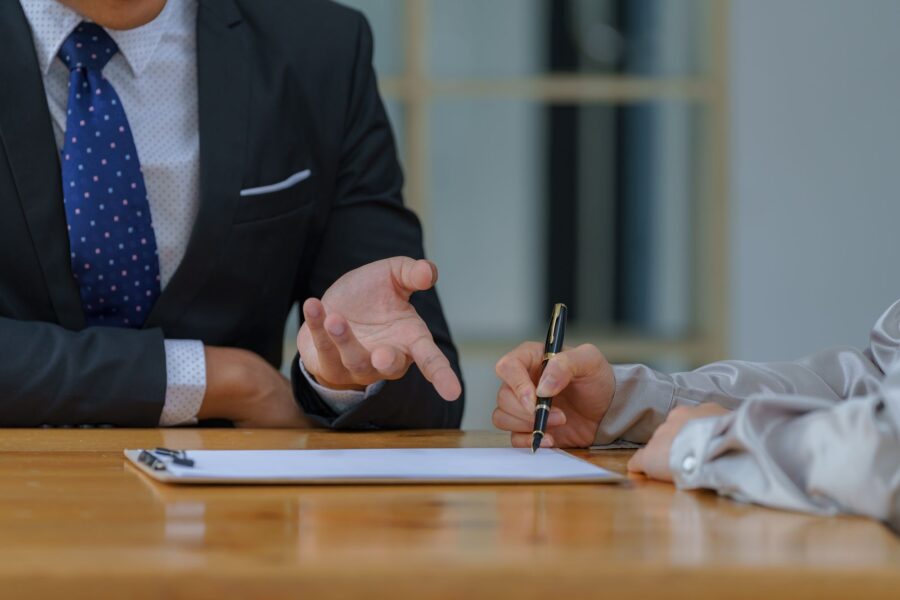 Two people at a meeting table, one gesturing while the other holds a pen over a document