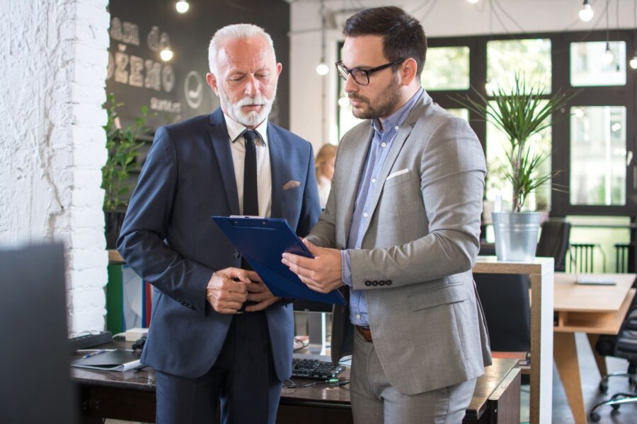 A senior and a younger executive in suits reviewing a document on a clipboard together in an office