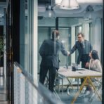 Two executives shake hands across a table in a glass-walled meeting room while a colleague looks on