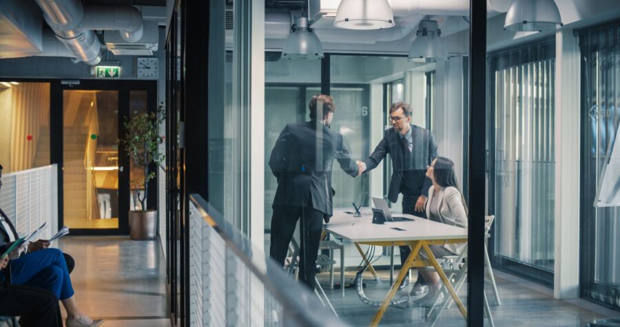 Two executives shake hands across a table in a glass-walled meeting room while a colleague looks on