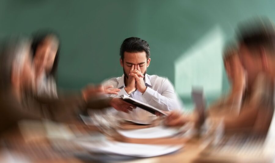 Executive with hands pressed together in frustration at a meeting table while colleagues talk around him